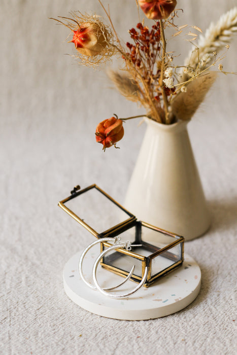 A small open jewellery box with a brass frame, holding two silver hoop earrings, styled on a round white coaster with dried flowers in a cream vase behind it.