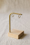 A pair of textured silver hoop earrings displayed on a round brass stand on a beige linen background.