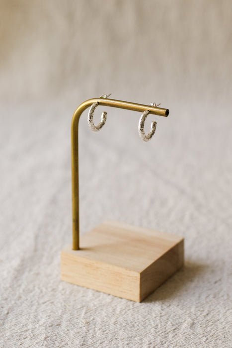 A pair of textured silver hoop earrings displayed on a round brass stand on a beige linen background.