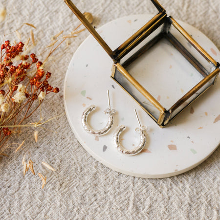 A pair of textured silver hoop earrings displayed on a round white coaster with a brass jewellery box, styled with dried flowers on a beige linen background.