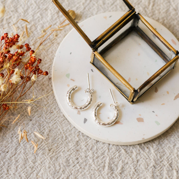 A pair of textured silver hoop earrings displayed on a round white coaster with a brass jewellery box, styled with dried flowers on a beige linen background.