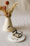 A pair of textured silver hoop earrings displayed on a round white coaster with a brass jewellery box, styled with dried flowers on a beige linen background.