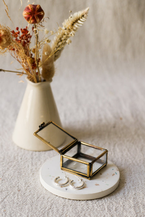 A pair of textured silver hoop earrings displayed on a round white coaster with a brass jewellery box, styled with dried flowers on a beige linen background.
