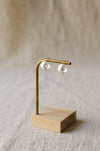 A pair of textured, hammered silver hoop earrings hanging from a brass display stand against a soft, beige linen background.