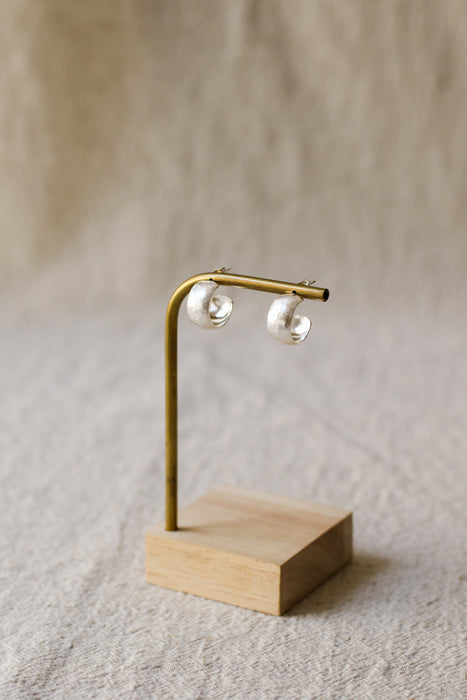 A pair of textured, hammered silver hoop earrings hanging from a brass display stand against a soft, beige linen background.