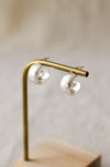 A pair of textured, hammered silver hoop earrings hanging from a brass display stand against a soft, beige linen background.