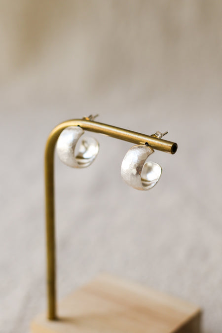 A pair of textured, hammered silver hoop earrings hanging from a brass display stand against a soft, beige linen background.