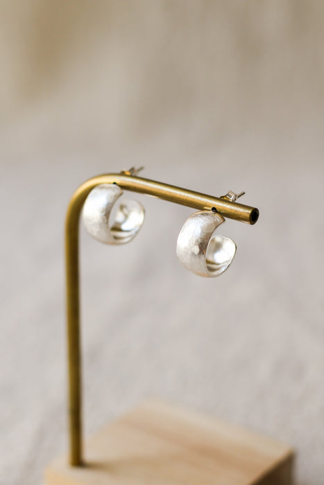 A pair of textured, hammered silver hoop earrings hanging from a brass display stand against a soft, beige linen background.