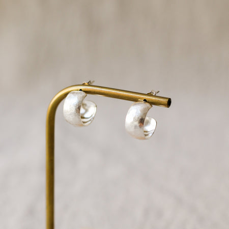 A pair of textured, hammered silver hoop earrings hanging from a brass display stand against a soft, beige linen background.
