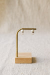 A pair of textured, hammered silver hoop earrings hanging from a brass display stand against a soft, beige linen background.