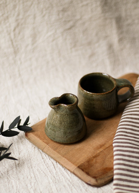 Green ceramic cup and milk jug on a wooden board with a striped cloth and leaves in the background.