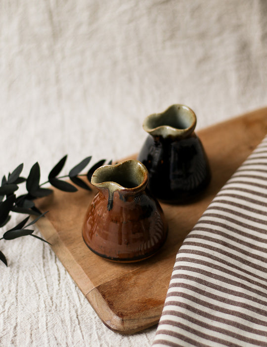 Two ceramic jugs on a wooden board with a striped fabric and greenery in the background.