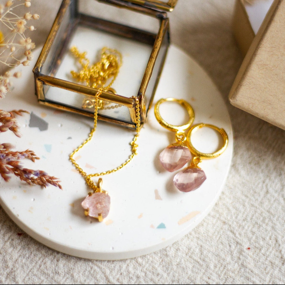 Gold hoop earrings and a gold chain necklace with rose quartz stones sitting on a white coaster, with a small glass jewellery box and dried flowers, all on a beige linen surface