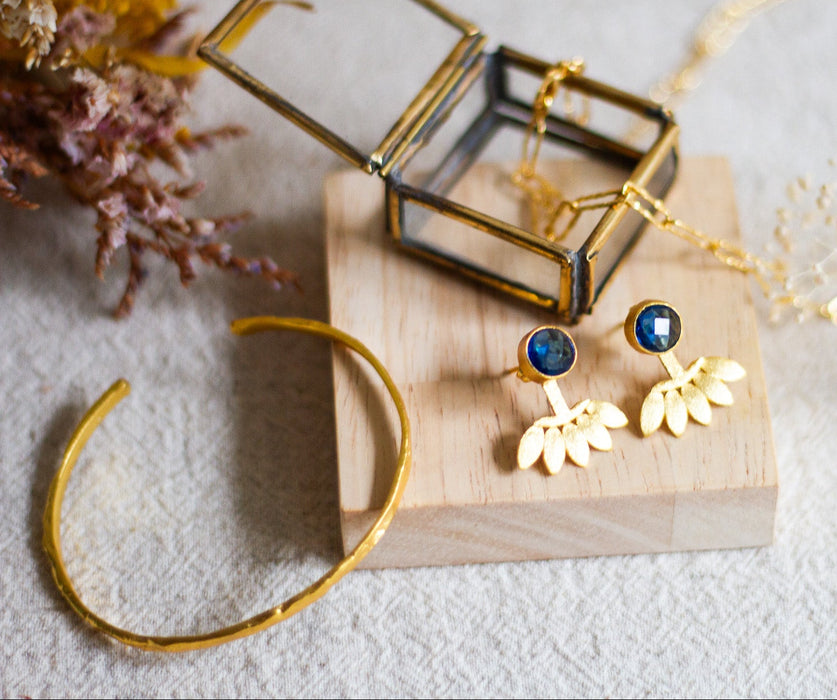 Gold jewellery set including stud earrings, bangle and a link necklace, on a wooden block with a glass jewellery box, on a wooden block with a linen background.
