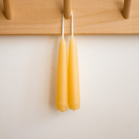Two beeswax candles hanging on a wooden peg rack against a white wall.