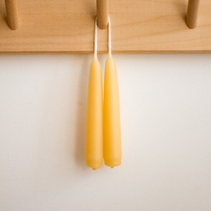 Two beeswax candles hanging on a wooden peg rack against a white wall.