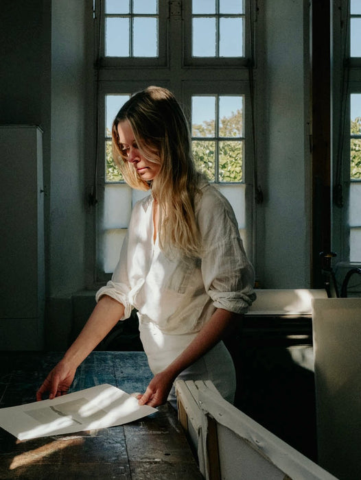 an artist in white clothes working on an etching in a studio with a wooden table, canvases and large windows in the background