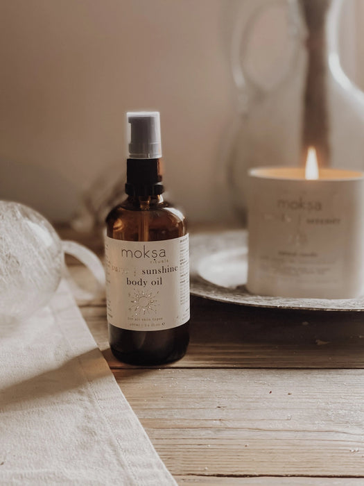 Bottle of moksa sunshine body oil on a wooden surface with a candle in the background.