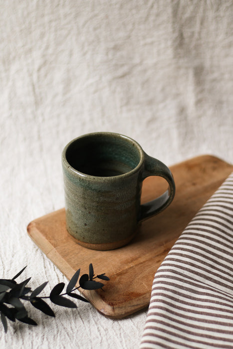 Green ceramic mug on a wooden board with a striped towel and leaves in the background