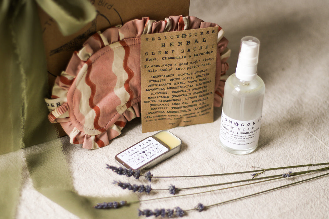 Herbal sleep sachet, bottle of pillow mist, pot of sleep balm, pink sleep mask, and lavender flowers on a beige linen surface.
