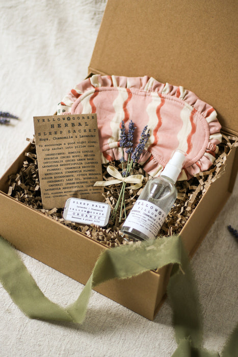 Gift set with pink sleep mask, bottle of pillow mist, sleep balm and sleep sachet and a bunch of lavender in a brown box on a linen surface.