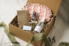 Kraft gift box containing a bottle of pillow mist, lavender flowers, and a pink fabric sleep mask, pot of sleep balm and brown sachet on a beige linen background with sage green ribbon and lavender 