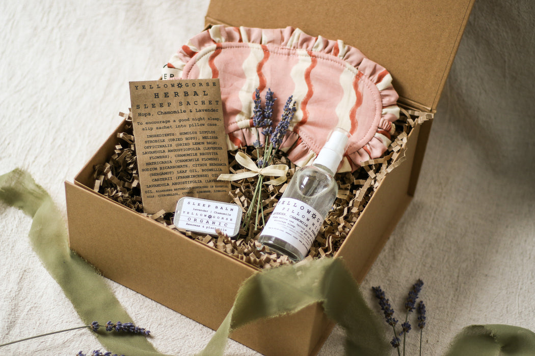 Kraft gift box containing a bottle of pillow mist, lavender flowers, and a pink fabric sleep mask, pot of sleep balm and brown sachet on a beige linen background with sage green ribbon and lavender 