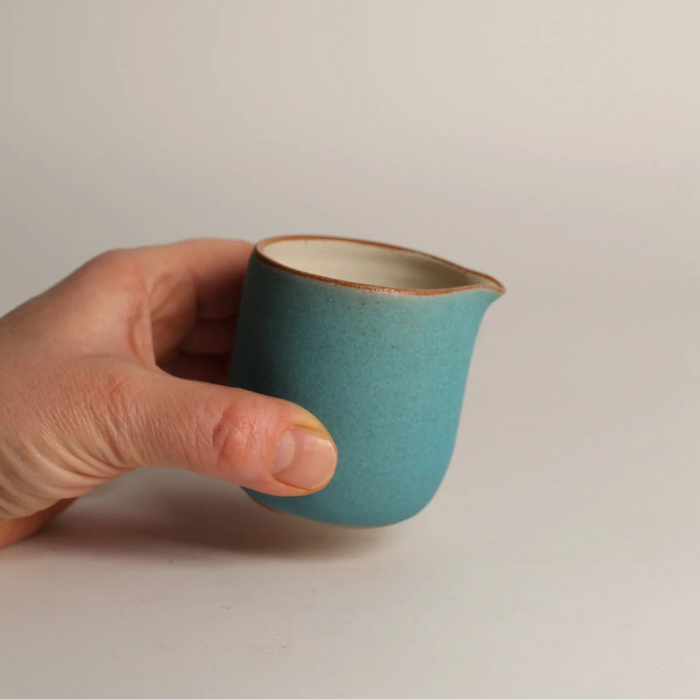 Hand holding a small turquoise ceramic jug against a plain grey background