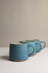 three matte turquoise stoneware coffee mugs on a neutral background.