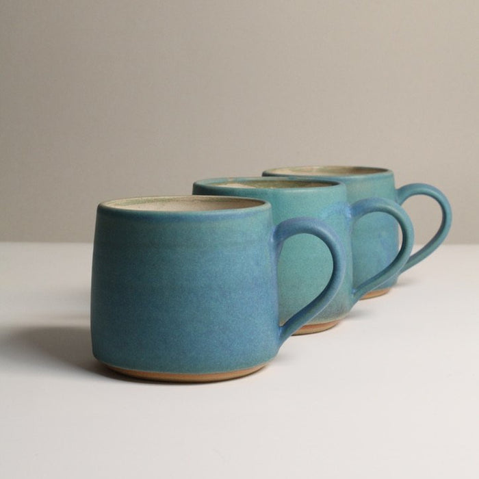 three matte turquoise stoneware coffee mugs on a neutral background.