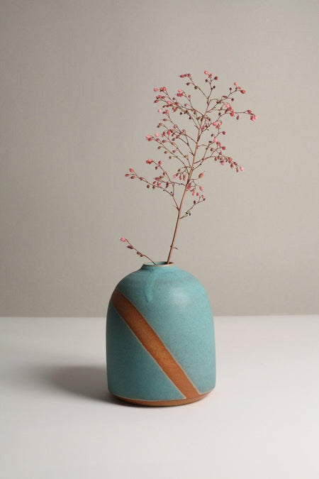 A matt turquoise ceramic vase with a brown stripe, containing a small branch with red berries, on a neutral background.