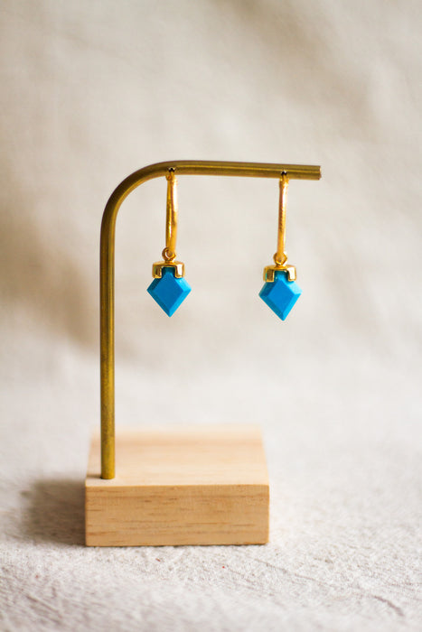Gold hoop earrings with turquoise stones on a brass and wooden stand against a neutral background