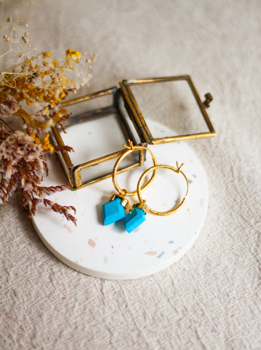 Gold hoop earrings with turquoise stones on a white round surface with dried flowers.