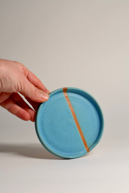 A hand holding a turquoise ceramic spoon rest with a visible brown stripe, against a white background