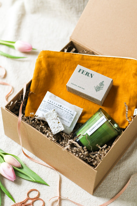 Gift box with a candle, soap, and other self care items, surrounded by pink tulips and pink ribbon, on a beige linen background 