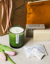 Green candle, soap bar, and pink tulips on a beige linen surface with a gift box in the background
