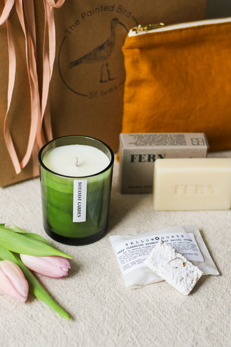 Green candle, soap bar, and pink tulips on a beige linen surface with a gift box in the background