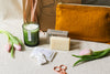 Green candle, soap, and linen pouch on a beige linen surface with tulips and sewing scissors