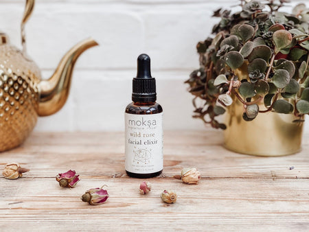 A bottle of Wild Rose Facial Elixir by Moksa, with a dropper, on a wooden surface surrounded by a potted plant and decorative items.
