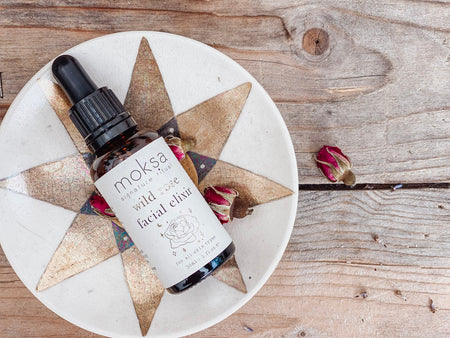 A bottle of Wild Rose Facial Elixir by Moksa, with a dropper, on a coaster and wooden surface.