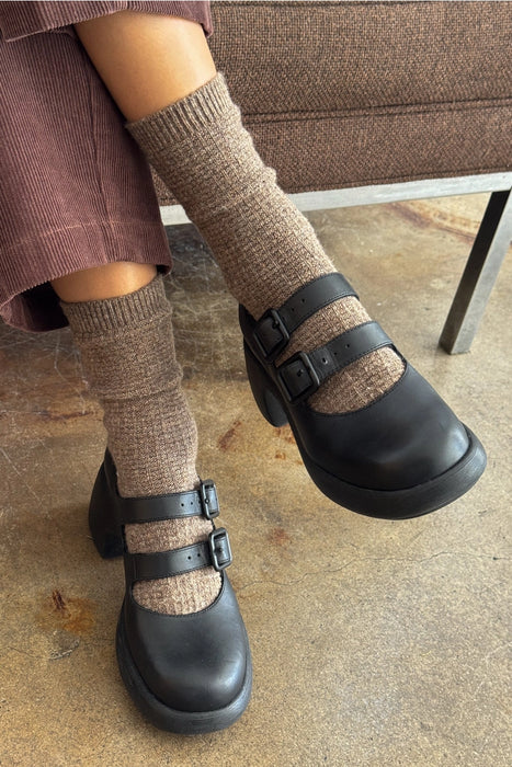 Person wearing black clogs with brown socks on a concrete floor.