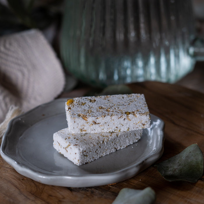 Two blocks of skincare scrub with yellow specks on a grey plate, with a blurred green vase and leaves in the background.