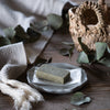 Green clay facial bar on a white plate with a woven basket and eucalyptus leaves in the background.