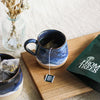 Hand thrown blue stoneware mug of tea, with a matching bowl filled with tea bags, and a vase of flowers, all on a wooden bread board, and beige linen tablecloth