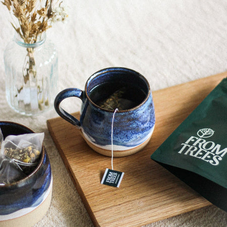 Hand thrown blue stoneware mug of tea, with a matching bowl filled with tea bags, and a vase of flowers, all on a wooden bread board, and beige linen tablecloth