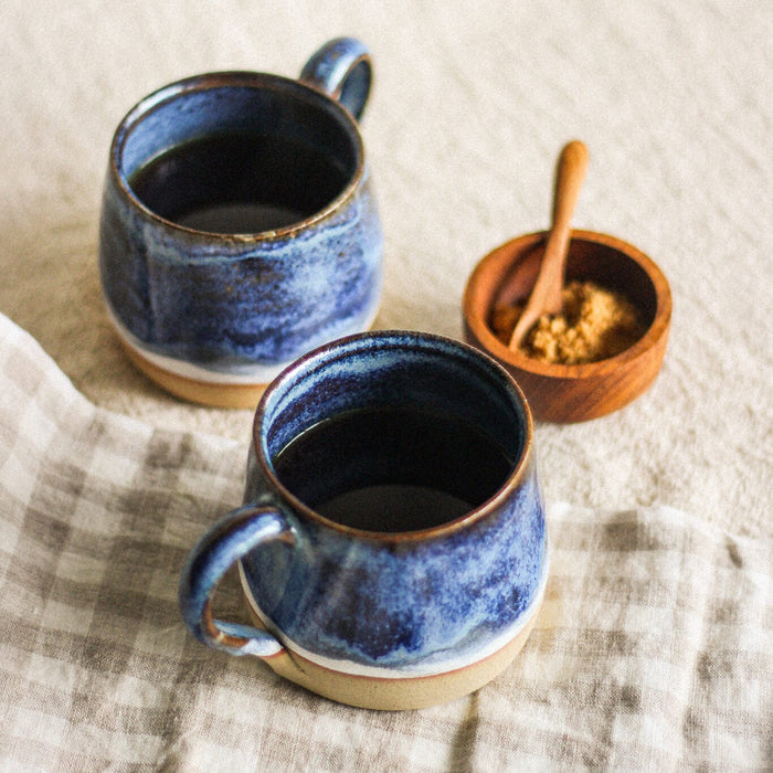 Two blue ceramic mugs on a textured surface with a small wooden bowl and spoon.