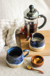 A hand thrown stoneware mug of coffee in a blue glaze, a matching milk jug, a wooden sugar pot and spoon, and a cafetière of coffee, on a  on a beige gingham surface and beige linen tablecloth
