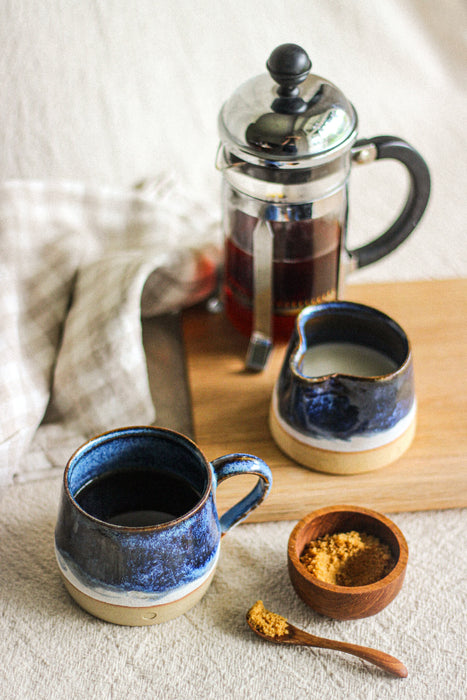 A hand thrown stoneware mug of coffee in a blue glaze, a matching milk jug, a wooden sugar pot and spoon, and a cafetière of coffee, on a  on a beige gingham surface and beige linen tablecloth