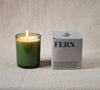 Lit candle in a green glass vessel next to a grey box labeled 'Fern' on a neutral linen background