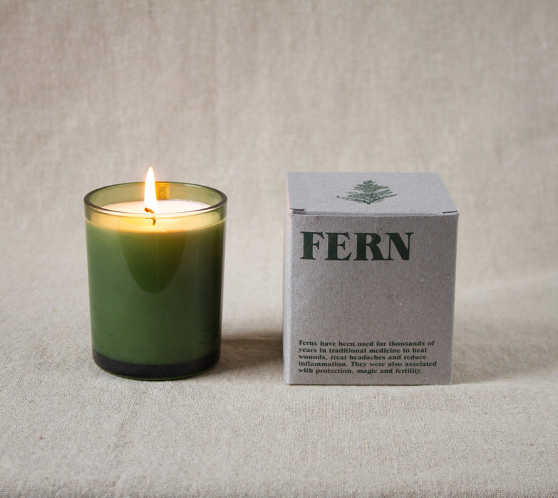 Lit candle in a green glass vessel next to a grey box labeled 'Fern' on a neutral linen background